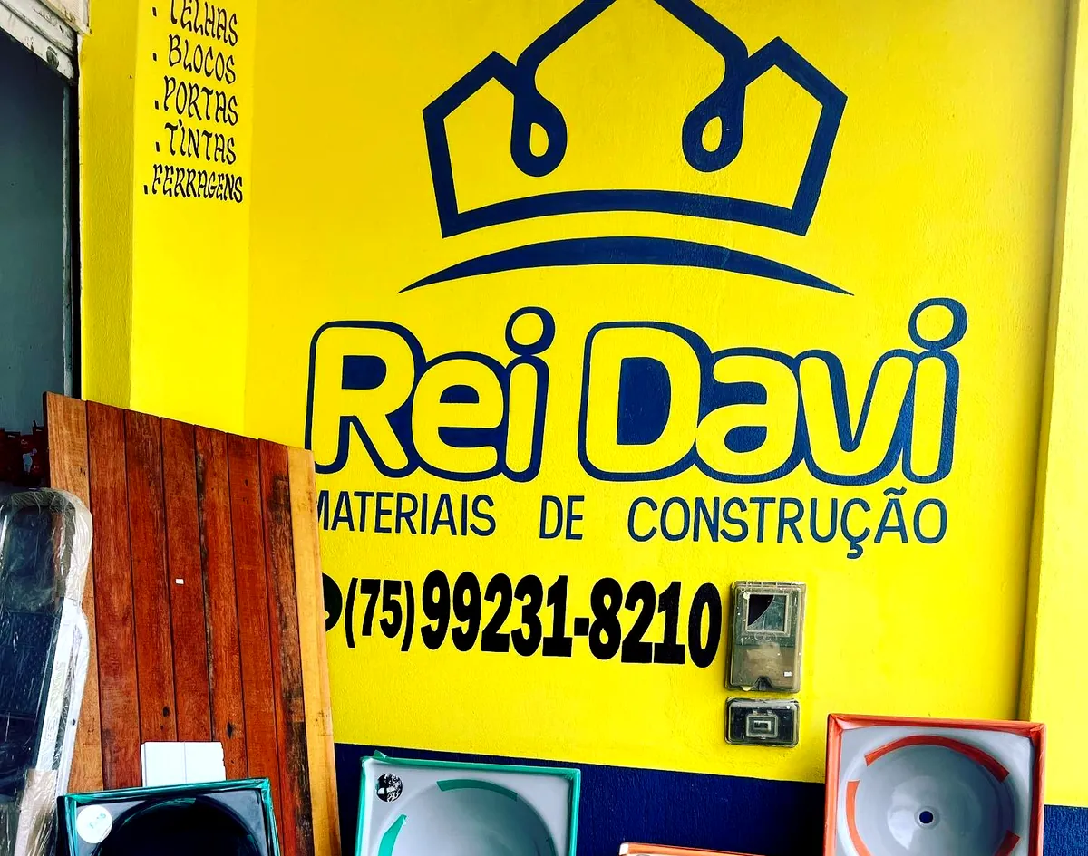 Parede amarela da Rei Davi Materiais de Construção com a logomarca da coroa, o WhatsApp da loja e tanques expostos na frente